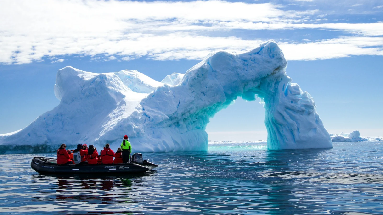 Expedition cruise passengers in red parkas on a Zodiac beside an iceberg arch