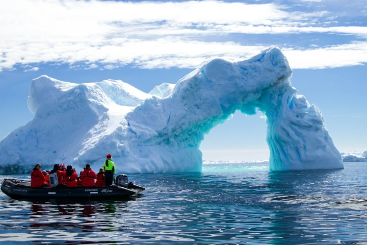 Expedition cruise passengers in red parkas on a Zodiac beside an iceberg arch