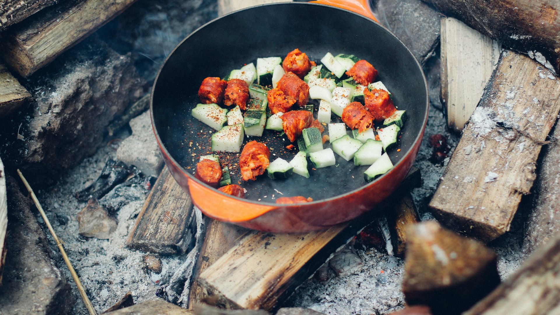 Vegetables and sausage cooking in a skillet over campfire coals