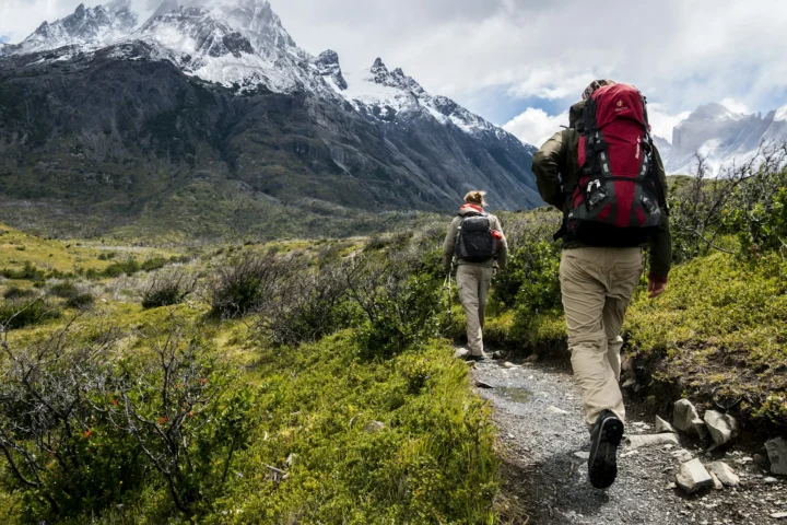 Two hikers with backpacks on a trail toward snowy peaks