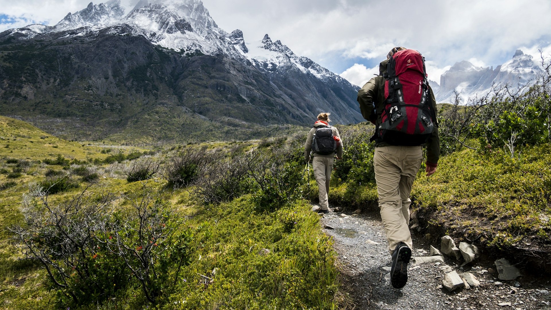 Two hikers with backpacks on a trail toward snowy peaks