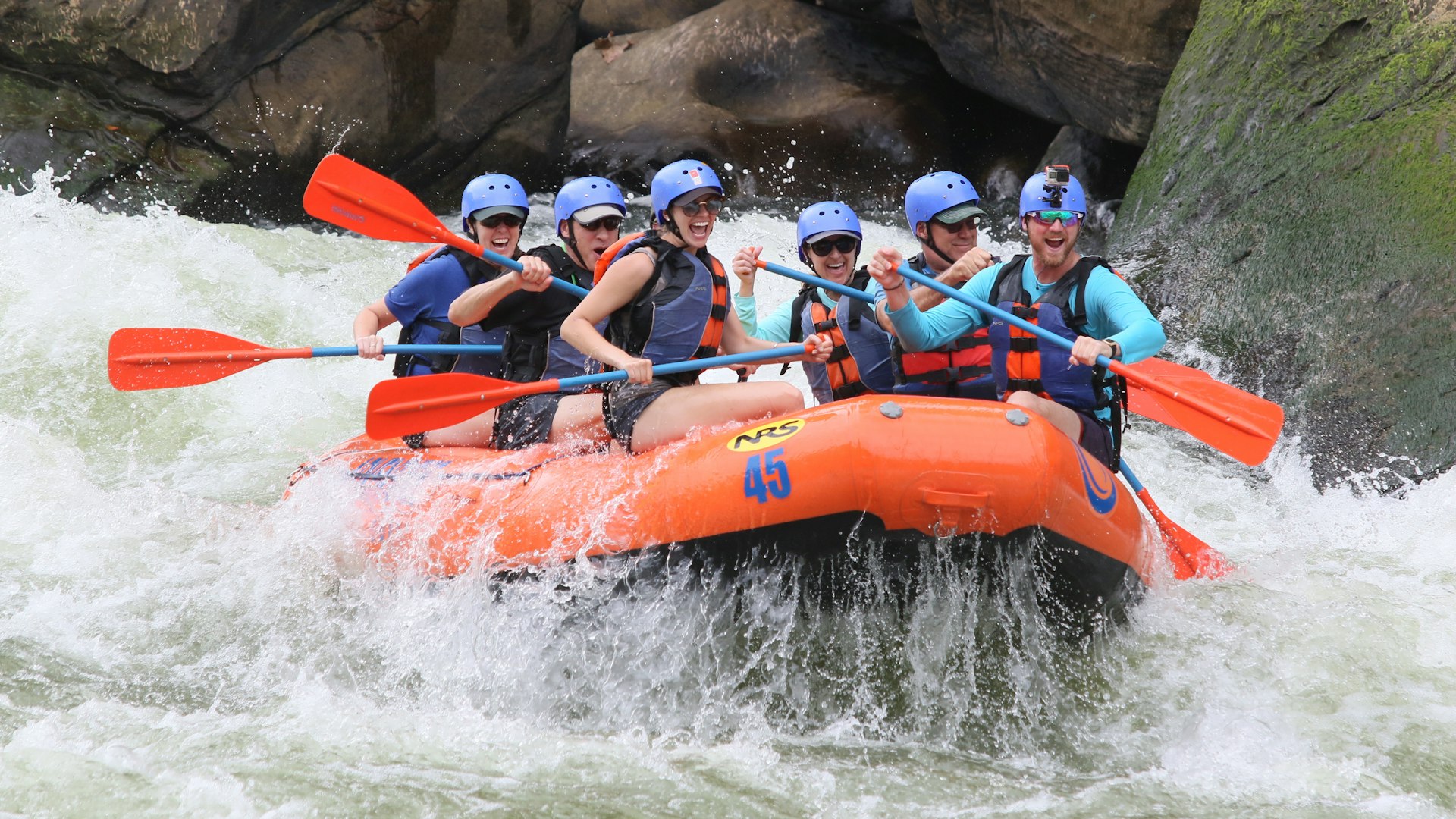 Rafting crew in helmets paddling an orange raft through whitewater